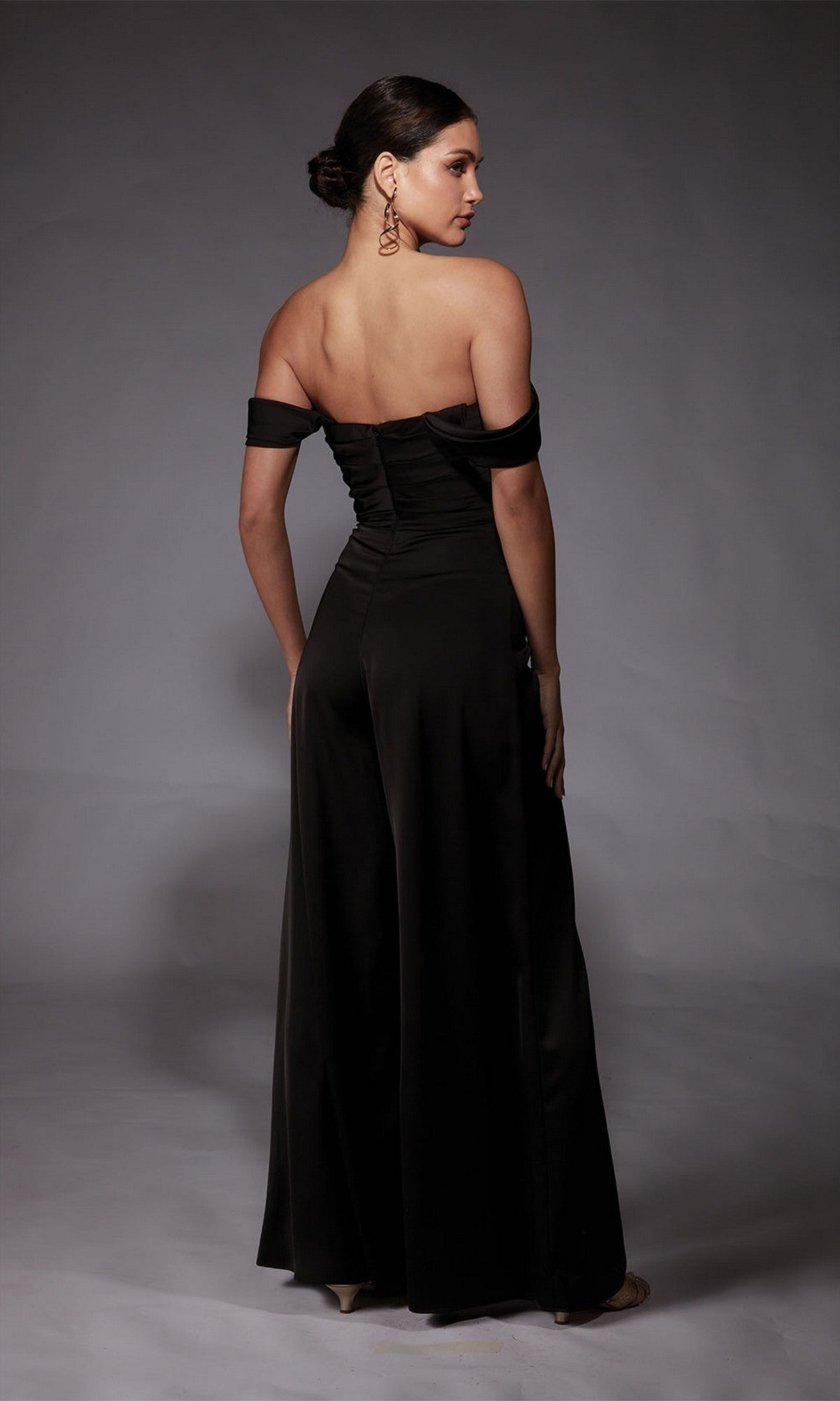 A woman faces away, wearing the Alyce 70071 Long Formal Jumpsuit with off-the-shoulder satin top, wide-leg pants, large earrings, and her hair in a low bun, set against a gray background.